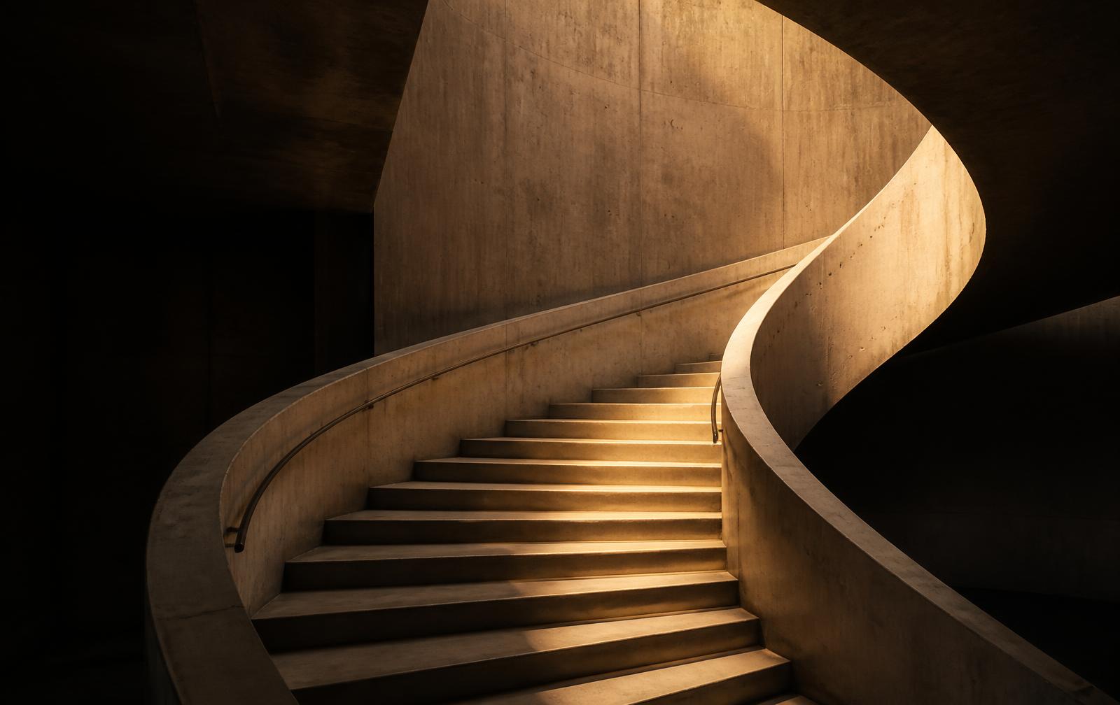A curving concrete spiral staircase lit by warm amber light rising out of deep charcoal shadow